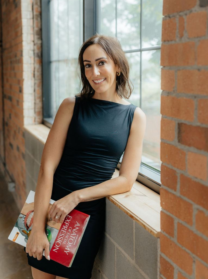 A woman in a black sleeveless dress stands by a large window with brick walls, smiling confidently. She holds a stack of magazines in her left arm. The natural light highlights her features, creating a warm and inviting atmosphere. This image conveys a sense of professionalism and approachability, making it suitable for lifestyle or fashion-related content.