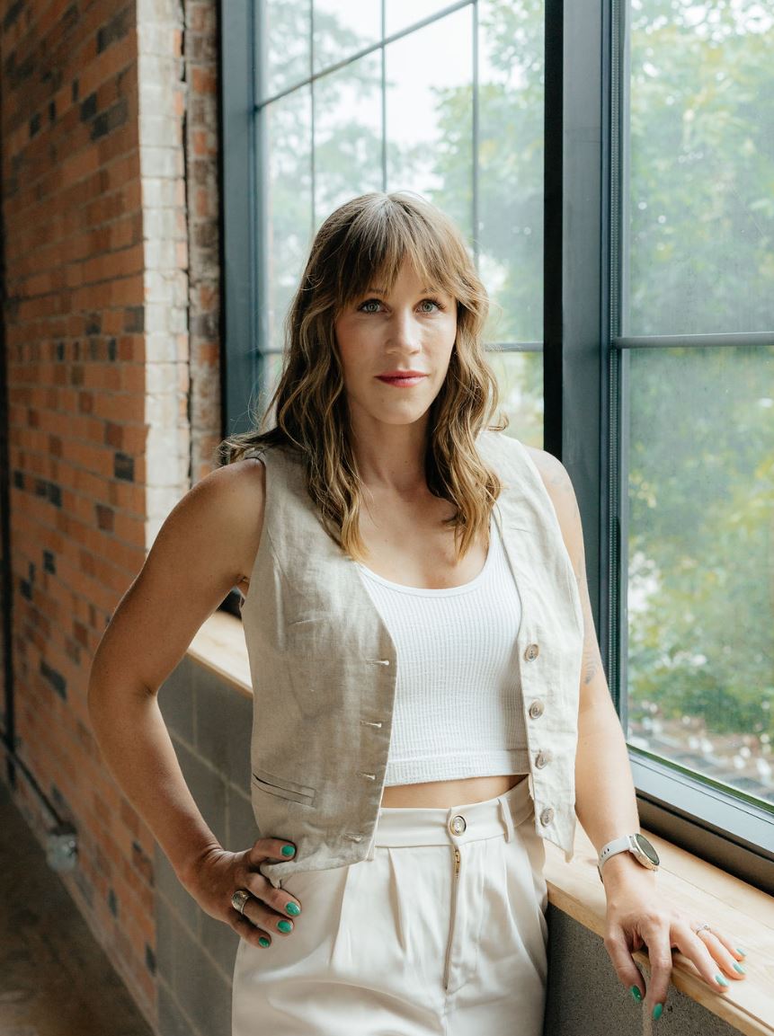 A woman stands confidently by a large window with natural light streaming in. She has long, wavy hair and wears a light-colored, sleeveless vest over a white top, paired with matching bottoms. The background features exposed brick walls, adding a modern yet rustic ambiance. This image captures a stylish, contemporary look, highlighting fashion and natural beauty in an indoor setting.