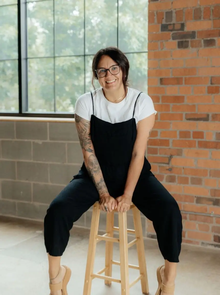 A person with glasses and tattoos sits on a wooden stool in a well-lit room featuring brick walls. They are wearing a white t-shirt underneath a black overall. The individual is smiling and positioned with their legs apart, creating a relaxed and approachable demeanor. Natural light from the large window enhances the warm atmosphere of the space, making it inviting and stylish. This image conveys a sense of confidence and casual fashion, suitable for lifestyle or fashion-related content.