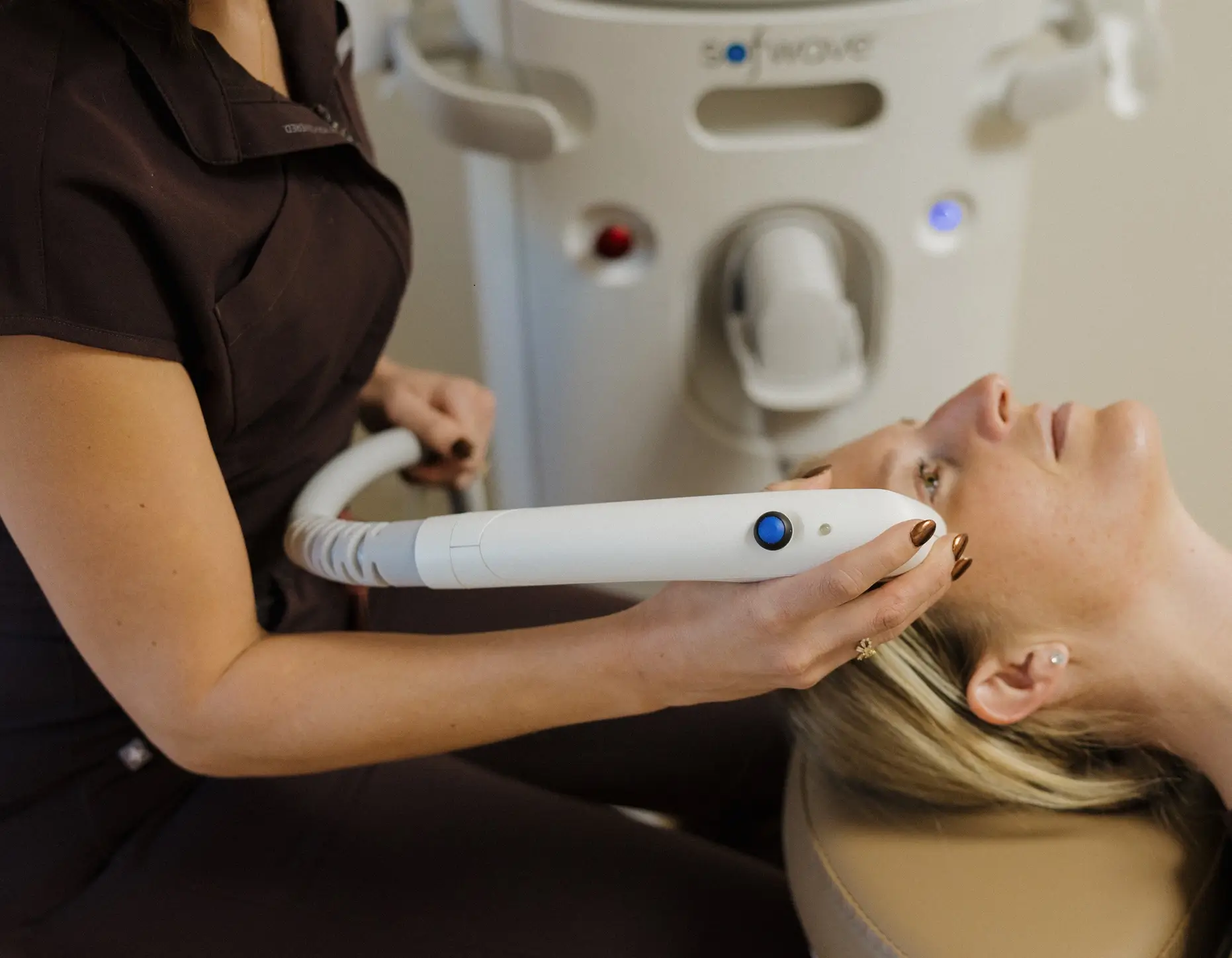 A skincare professional performs a facial treatment using a handheld device on a client lying on a treatment table. The setting is a modern spa or clinic, featuring advanced skincare technology in the background. The procedure aims to enhance skin health and appearance, showcasing the focus on personalized skincare solutions. The image highlights the professional environment and the care given to clients during treatments.