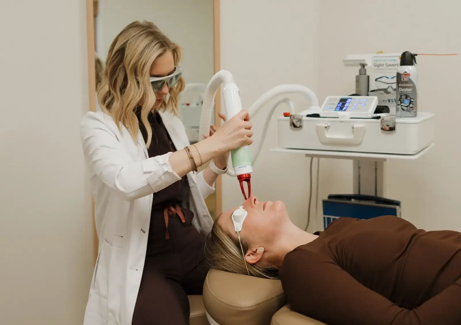A skincare professional wearing protective eyewear administers a laser treatment to a client lying on a treatment table in a modern clinic. The clinician uses a handheld laser device focused on the client's facial area, aimed at enhancing skin health and appearance. The clinic is equipped with various medical instruments and supplies, reflecting a clean and professional environment. This image illustrates advanced skincare procedures, emphasizing the importance of professional care in achieving optimal skin results.