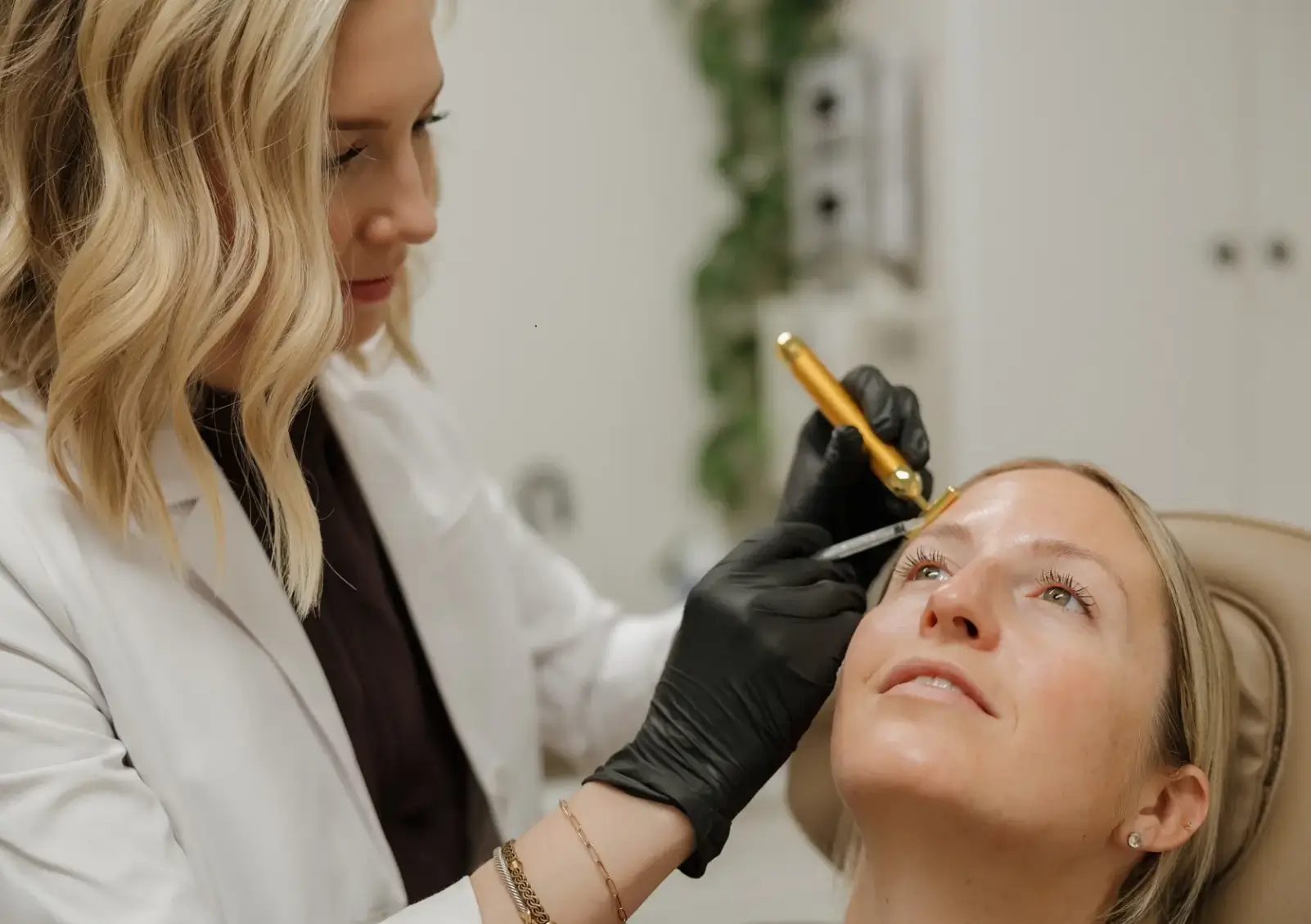 A licensed aesthetician performs a cosmetic procedure on a client in a modern beauty clinic. The aesthetician, wearing a white lab coat and black gloves, carefully uses a microneedling tool on the client's eyebrow area. The client appears relaxed and is seated comfortably, with a focus on enhancing her facial features. Soft, neutral-colored decor in the background creates a calming atmosphere. This image highlights the professional environment of aesthetic treatments, emphasizing safety, expertise, and client care.