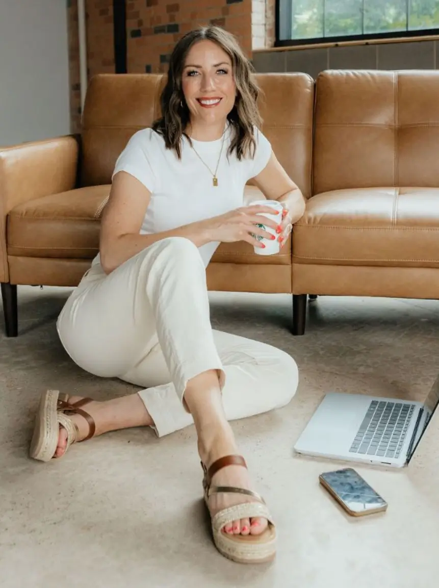 Young woman sitting on a carpeted floor in a cozy living room, wearing a white t-shirt and cream pants, holding a coffee cup, with a laptop and smartphone nearby, exuding a relaxed and productive atmosphere.