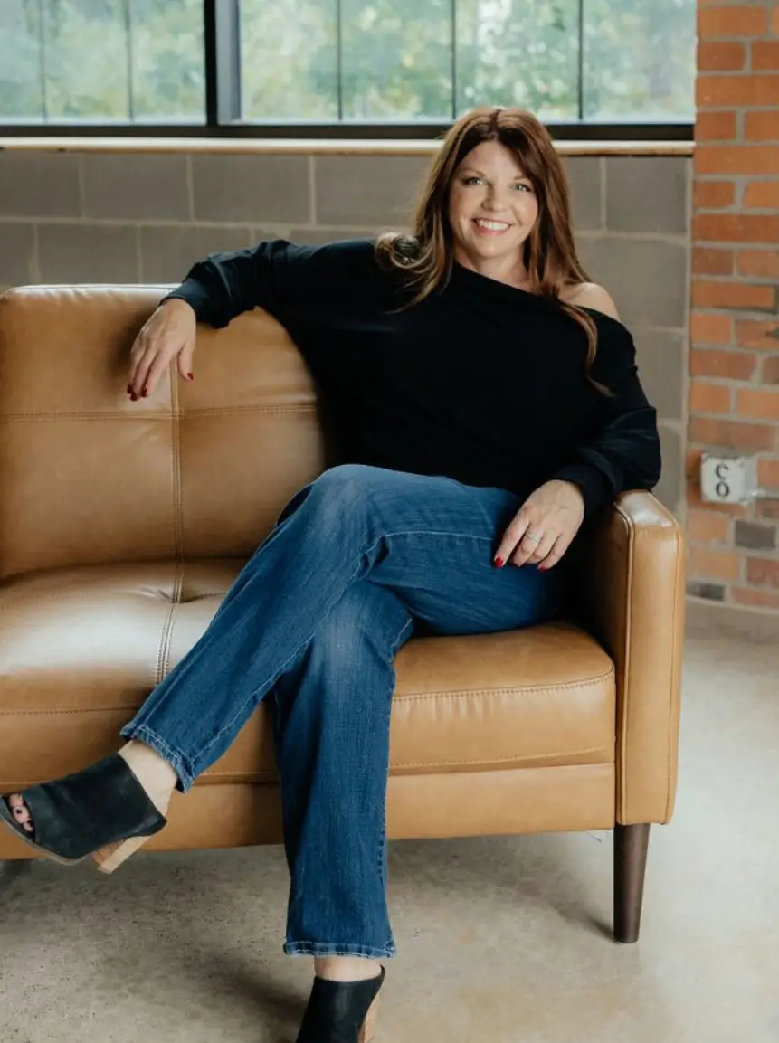 Woman in a black off-shoulder sweater and blue jeans sitting casually on a tan leather sofa, with a modern interior background.