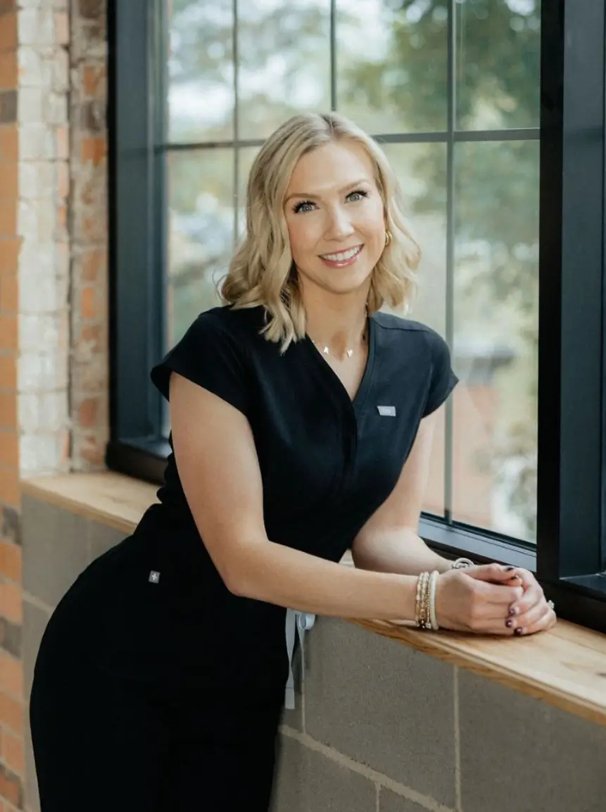 Professional woman in a black dress leaning against a wooden counter by a large window, smiling confidently.