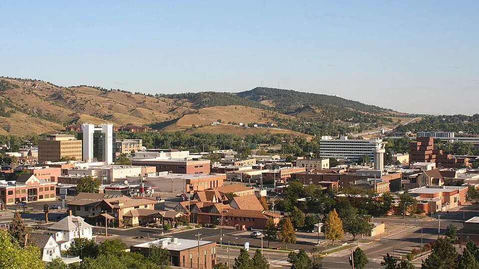 Panoramic view of downtown Rapid City, South Dakota, with city buildings and the Black Hills in the background.